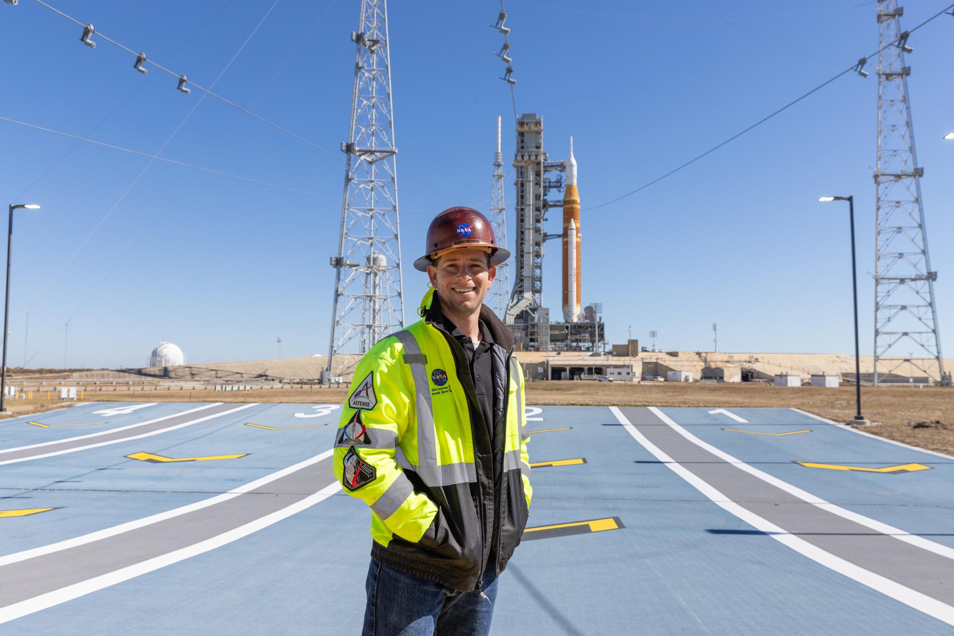 Image shows Jesse Berdis standing standing at the pad of Launch Complex 39B at NASA's Kennedy Space Center in Florida. Behind him are clear blue skies and NASA's SLS (Space Launch System) rocket and Orion spacecraft for the Artemis II mission. Photo credit: Kim Shiflett